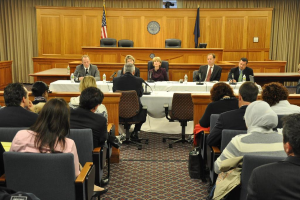Ein Versammlungsraum mit einer Gruppe von Menschen, die auf Stühlen sitzen, zwei fremde Flaggen, ein oranger Schreibtisch mit Wasserflaschen und einem Mikrofon.