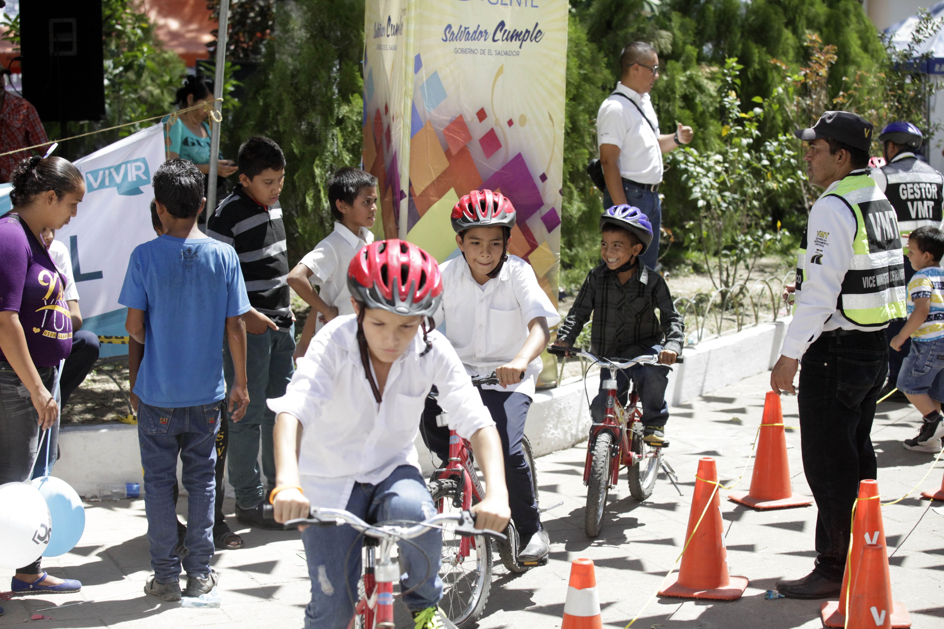 Kinder fahren Fahrräder auf einer Straße mit Verkehrsleitkegeln, einige tragen Helme, andere stehen daneben, mit einem Banner, Bäumen und Gebäuden im Hintergrund.