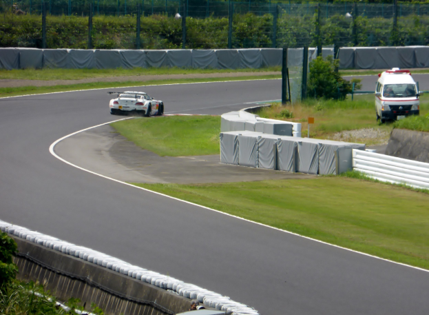 Ein Rennauto, das auf einer Rennstrecke fährt, mit einem Polizeiwagen im Hintergrund, umgeben von Gras, Pflanzen, Bäumen und einem Fahrzeug auf der rechten Seite und Text unten.