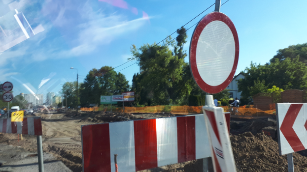 Baustelle mit einem roten und weißen Schild am Straßenrand, Bäume, Straßenlaternen, ein Verkehrszeichen, Gebäude und einen klaren blauen Himmel im Hintergrund.