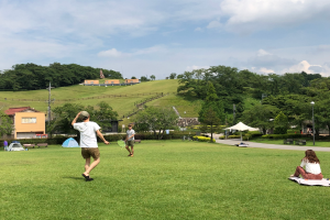 Eine Gruppe von Menschen, die Badminton in einem Park spielen, mit einem Mann und einer Frau auf einer Decke auf dem Rasen sitzend, der Mann hält einen Schläger in der Hand, und Zelte, Straßenlaternen, Strommasten, Stromkabel, Gebäude, Bäume, Hügel und ein bewölkter Himmel im Hintergrund.