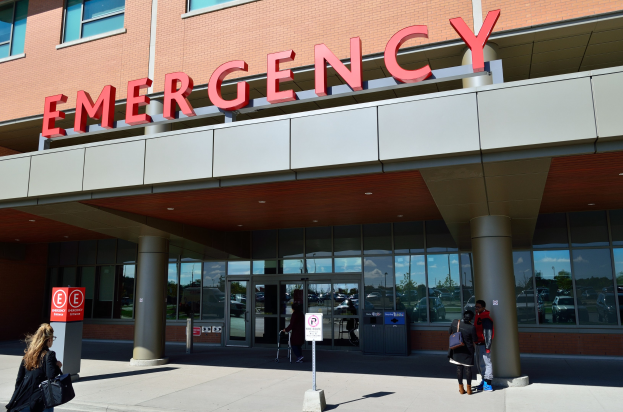 Gruppe von Menschen, die auf ein Notaufnahme-Gebäude mit Glaswänden, Säulen und einem Namensschild zugehen, neben Schildern und einem Müllcontainer.