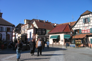 Gruppe von Menschen, die eine Kopfsteinpflasterstraße in einem Stadtplatz entlanggehen, mit Gebäuden, Schildern, Laternen, Pflanzen und Fahrzeugen, unter einem sichtbaren Himmel.