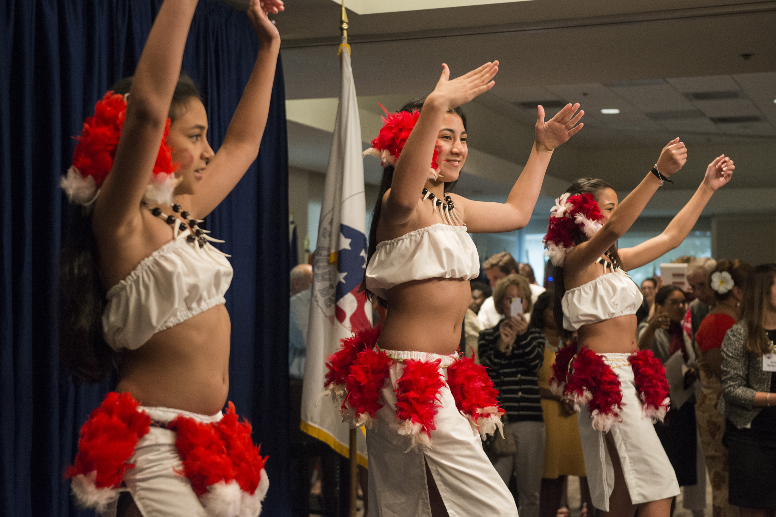 Eine Gruppe junger Frauen in weißen und roten Outfits tanzt vor einem Publikum, mit einer Fahne, Vorhängen und Deckenlampen im Hintergrund.
