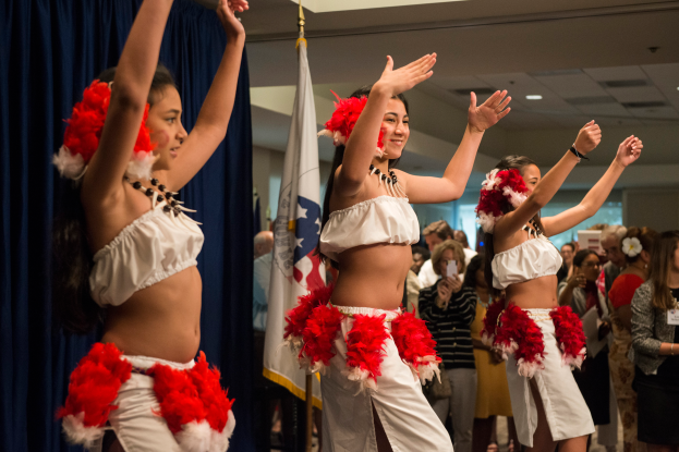 Eine Gruppe junger Frauen in weißen und roten Outfits tanzt vor einem Publikum, mit einer Fahne, Vorhängen und Deckenlampen im Hintergrund.