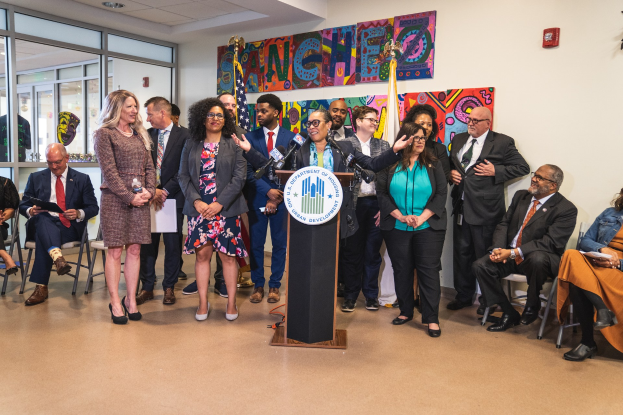 Gruppe von Menschen vor einem Rednerpult mit Mikrofon und einer Tafel mit der Aufschrift "Sancho Stadtverordnetenvorsteher tritt in die Stadtverordnetenversammlung ein", mit sitzenden und stehenden Teilnehmern, Flaggen, Feuerlöscher und Deckenleuchten im Hintergrund.