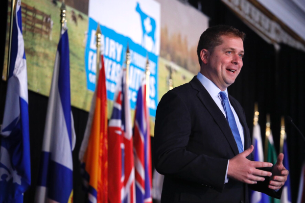 Ein Mann in Anzug und Krawatte steht an einem Rednerpult mit Mikrofon, umgeben von Fahnen und einem Bildschirm im Hintergrund, und scheint eine Äußerung zu machen.