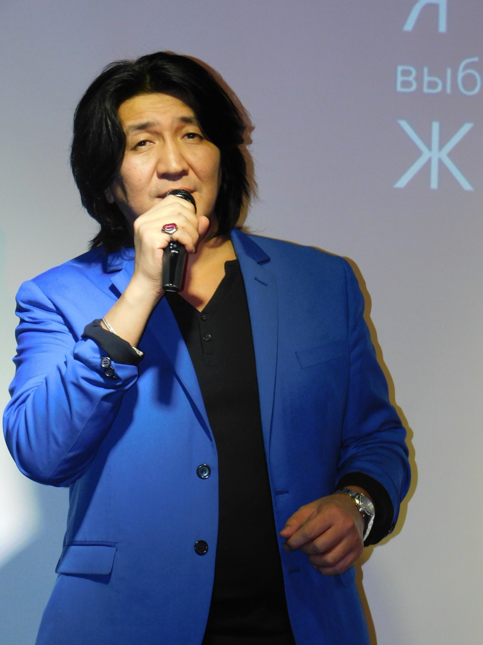 Ein Mann in einem blauen Anzug mit einem Mikrofon, der vor einem Bildschirm mit Text steht, trägt einen schwarzen Anzug und ein weißes Hemd, hat ordentlich gekämmtes Haar und einen ernsten Gesichtsausdruck.