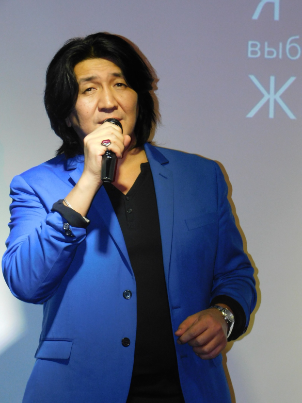 Ein Mann in einem blauen Anzug mit einem Mikrofon, der vor einem Bildschirm mit Text steht, trägt einen schwarzen Anzug und ein weißes Hemd, hat ordentlich gekämmtes Haar und einen ernsten Gesichtsausdruck.