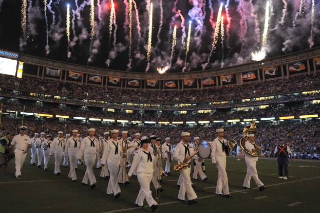 Eine Gruppe von Menschen in weißen Uniformen marschiert auf einem Fußballfeld, einige halten Musikinstrumente, während Zuschauer von den Tribünen zuschauen, Banner und Stadionbildschirme sind sichtbar und Feuerwerk erhellt den Himmel darüber.