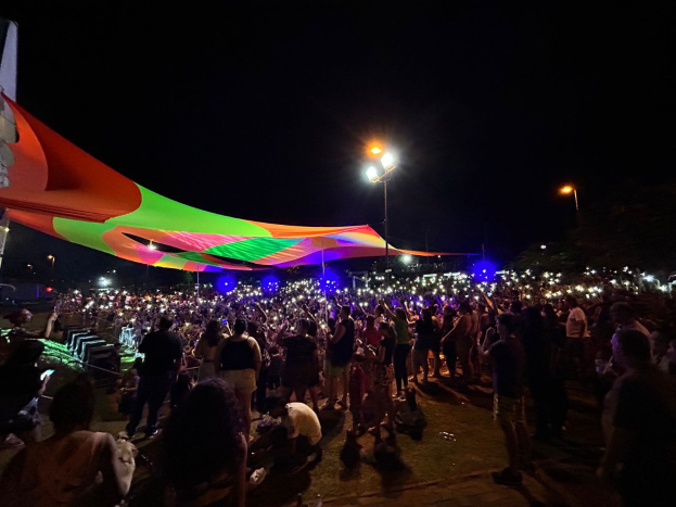 Eine große Menschenmenge steht vor einem hell erleuchteten Zelt bei Nacht während des 2018er Kunstfestivals, mit bunten Lichtern, beleuchteten Pfählen, einem Gebäude auf der linken Seite und dem Nachthimmel im Hintergrund.