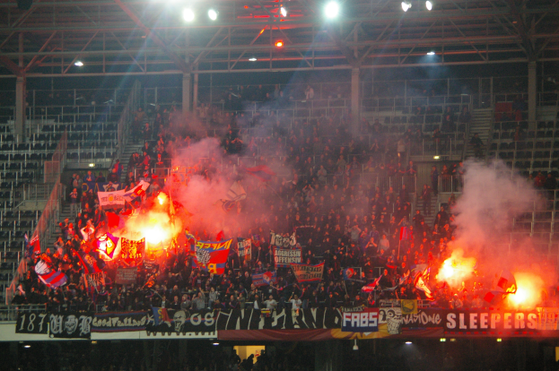 Eine große Menge Menschen in einem Stadion hält Fahnen und Banner, mit Leuchtfeuern, die Rauch erzeugen, unter einer Decke mit Deckenleuchten und Metallrahmen.