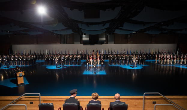 Eine große Gruppe uniformierter Personen steht auf einer Bühne mit einem Podium und einem Mikrofon, während unten sitzende Personen und eine beleuchtete Wand im Hintergrund zu sehen sind.