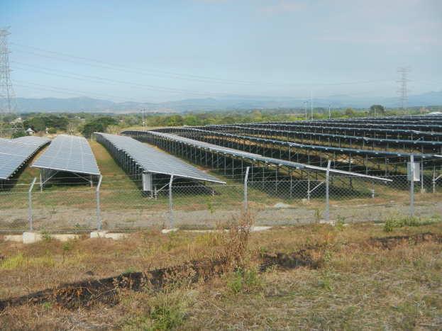 Ein Solarpanel-Feld, das von einem Zaun umgeben ist, mit Gras und Pflanzen im Vordergrund, Bäumen, Übertragungstürmen mit Drähten und Hügeln im Hintergrund unter einem klaren blauen Himmel.