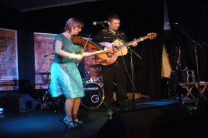 Ein Mann in schwarzem Hemd und Hose hält eine Gitarre in der Mitte der Bühne, während eine Frau in einem blauen Kleid links von ihm eine Geige spielt und verschiedene Musikinstrumente, darunter Lautsprecher und Trommeln, im Hintergrund zu sehen sind.