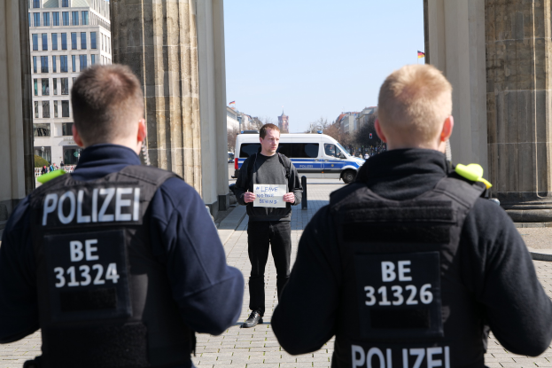 Drei Polizeibeamte stehen vor einem Gebäude, ein Mann hält ein Papier in der Hand, flankiert von Säulen, während Fahrzeuge, Menschen, Bäume und Gebäude im Hintergrund zu sehen sind.