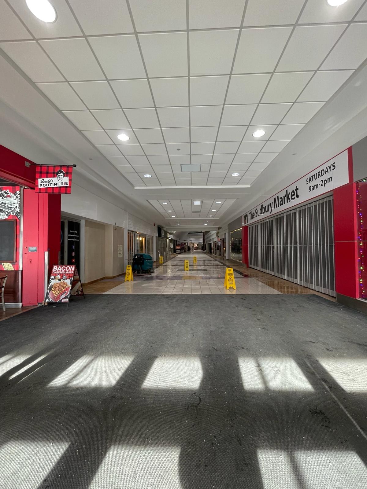 Innenansicht eines Einkaufszentrums mit Deckenbeleuchtung, einem Stuhl und einer Tafel mit der Aufschrift "Sydbury Market - Vorher und Nachher" links und verstreuten Gegenständen rechts und auf dem Boden.