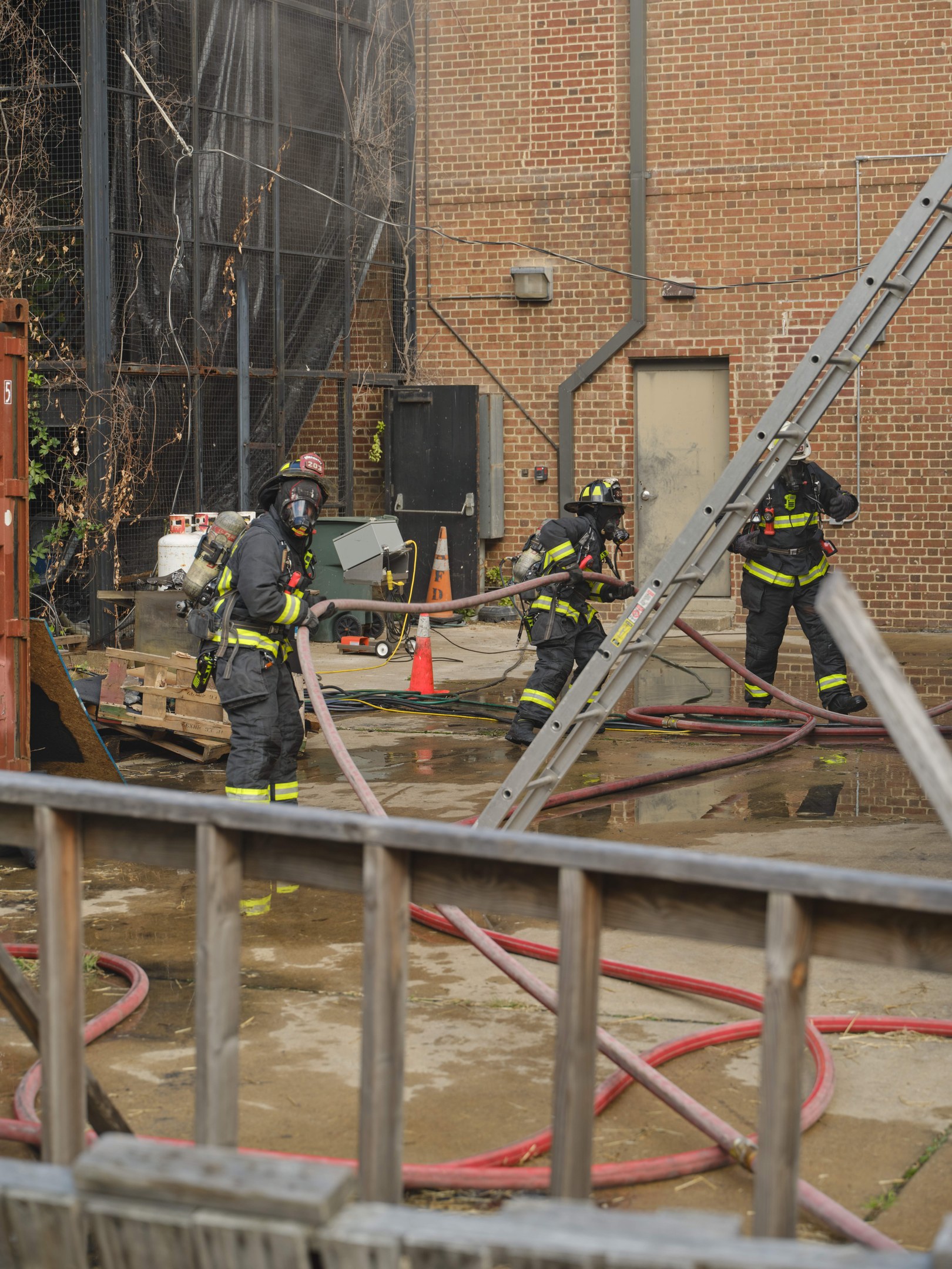 Feuerwehrleute mit Helmen arbeiten daran, ein Gebäude Feuer zu löschen, während sie Schläuche halten, mit einem Metallzaun, verstreuten Rohren, einem Container, einem Verkehrskegel und Hintergrundelementen einschließlich eines Baums und des Himmels.