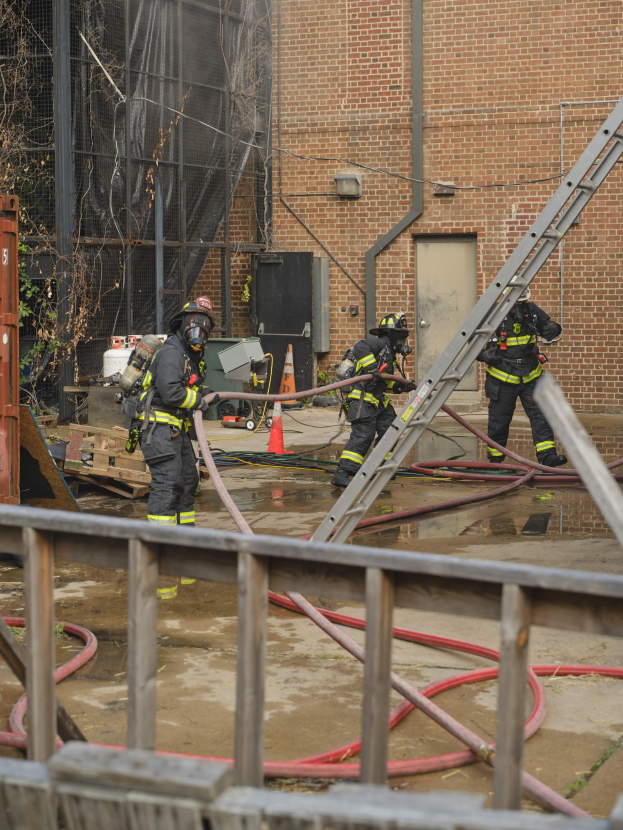 Feuerwehrleute mit Helmen arbeiten daran, ein Gebäude Feuer zu löschen, während sie Schläuche halten, mit einem Metallzaun, verstreuten Rohren, einem Container, einem Verkehrskegel und Hintergrundelementen einschließlich eines Baums und des Himmels.