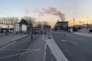 Eine Stadtstraße mit Verkehrskegeln, einem Bus, Laternen, Bäumen, Gebäuden, Kränen und Rauch, der aus einem fernen Gebäude aufsteigt.