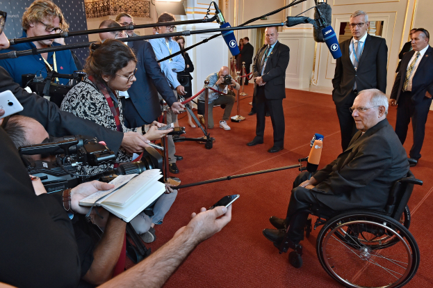 Ein Mann im Rollstuhl umgeben von Reportern mit Kameras, Mikrofonen und Handys, mit einem Banner und Scheinwerfern im Hintergrund.