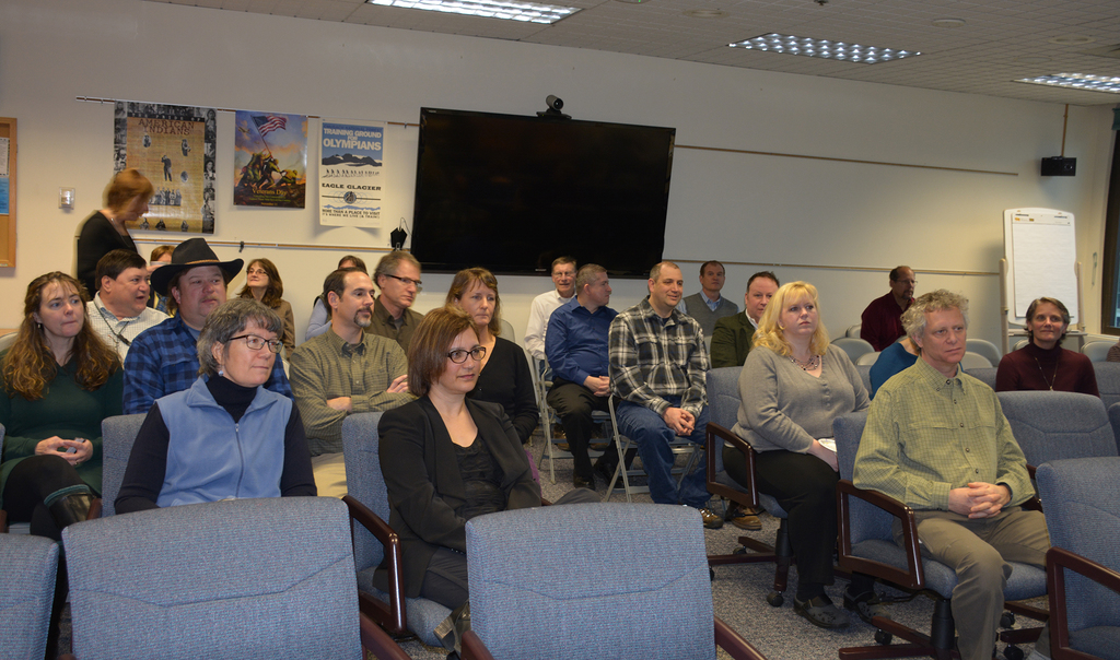 Eine Gruppe von Menschen, sowohl Männer als auch Frauen, sitzt auf Stühlen in einem Raum mit einem Fernseher, Papieren an der Wand und einer Tafel auf der rechten Seite.