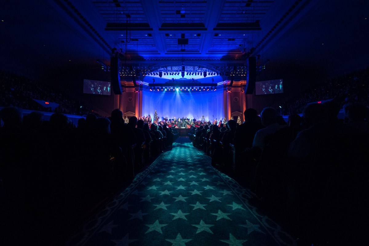 Großer Saal mit sitzendem Publikum, Bühne mit Musikern und Deckenleuchten und -fernsehern, typisch für einen Konzertsaal der Nationalen Convention 2019.