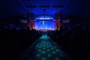 Großer Saal mit sitzendem Publikum, Bühne mit Musikern und Deckenleuchten und -fernsehern, typisch für einen Konzertsaal der Nationalen Convention 2019.