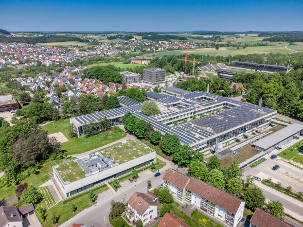 Luftaufnahme eines großen Gebäudes mit Solarpaneelen auf dem Dach, umgeben von Häusern, Bäumen, Fahrzeugen, Hügeln und einem klaren blauen Himmel, identifiziert als Hauptsitz eines deutschen Solarenergieunternehmens.
