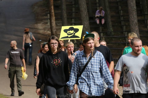 Gruppe von Menschen, die einen Waldweg entlanggehen, einige tragen Taschen und einer hält ein Plakat mit einem Personenbild, mit Bäumen und Treppen im Hintergrund, wahrscheinlich an einer Demonstration teilnehmend.