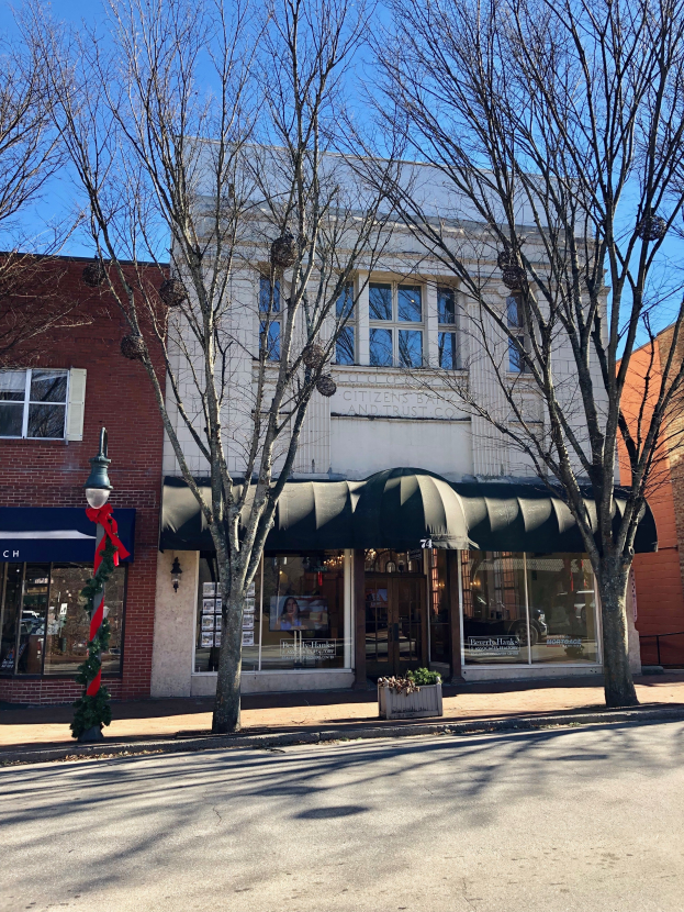 Ein Eckgebäude mit einer grünen Markise, Glasfenstern und einem Namenbrett, auf dem 'Citizens Bank and Trust Co.' steht, umgeben von Bäumen und einem Laternenmast, mit einem bewölkten Himmel im Hintergrund.