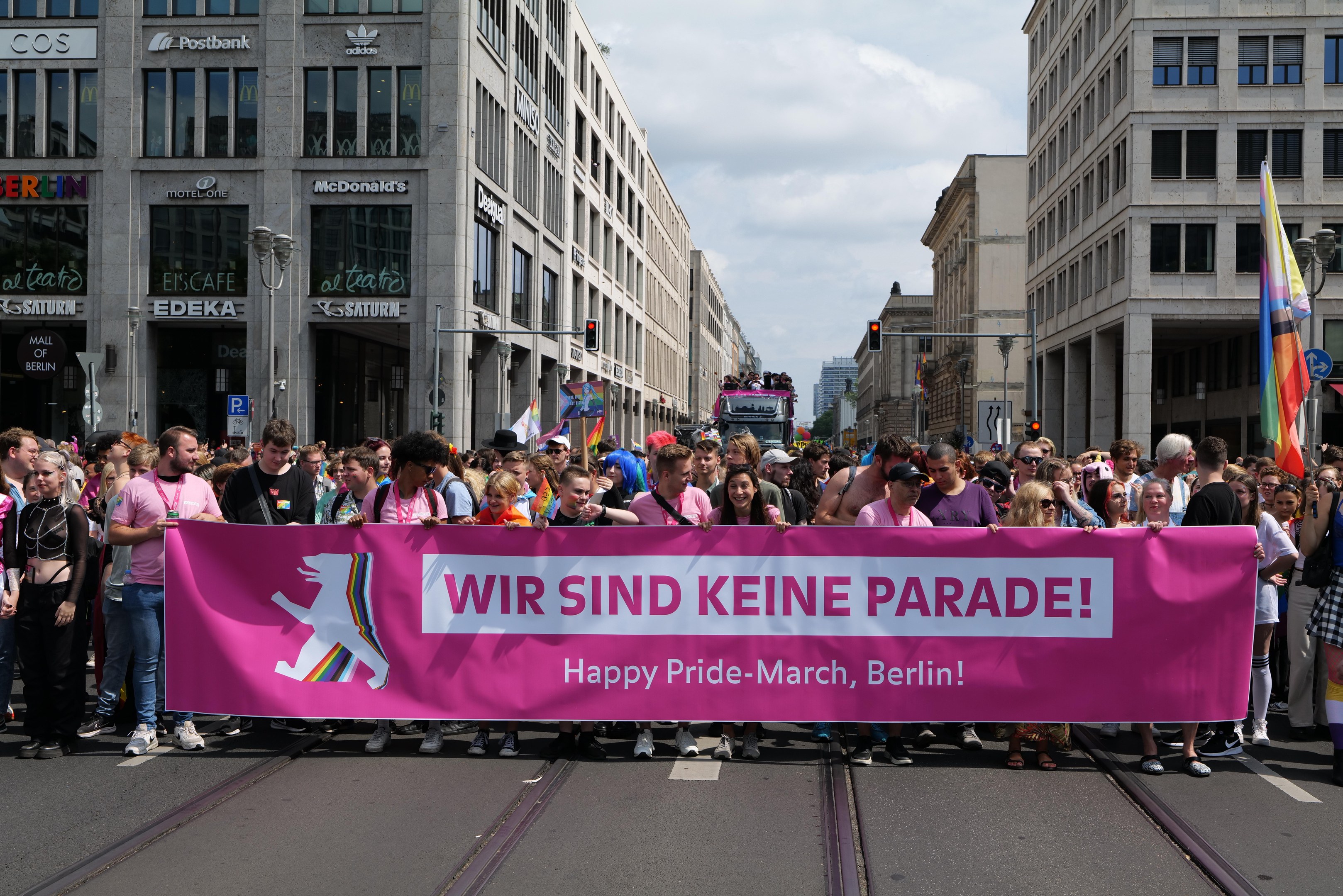 Eine Gruppe von Menschen marschiert auf einer Straße in Berlin mit einem pinken Banner, auf dem "Happy Pride March" steht, vorbei an Gebäuden, Laternenmasten und Verkehrszeichen.
