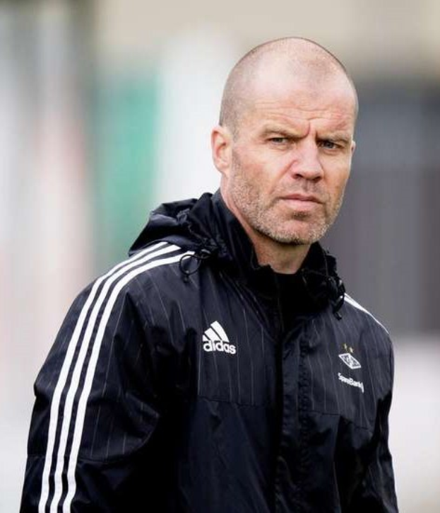 Mann in marineblauer Jacke mit weißen Streifen und weißem Kragen, der Hände verschränkt, auf einem Fußballfeld mit ernstem Gesichtsausdruck und unscharfem Hintergrund steht.