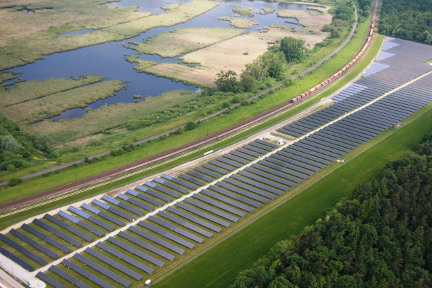 Luftaufnahme einer Solaranlage mit Panelen in einem Feld, umgeben von Bäumen, Gras, Pflanzen und Wasser, mit einem Zug auf einer nahen Bahnstrecke.