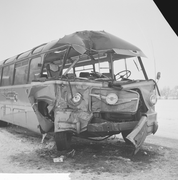 Schwarz-weiß-Foto eines schwer beschädigten Busses mit eingedellter Front und zersplitterten Fenstern am Straßenrand, umgeben von Bäumen unter einem bewölkten Himmel.