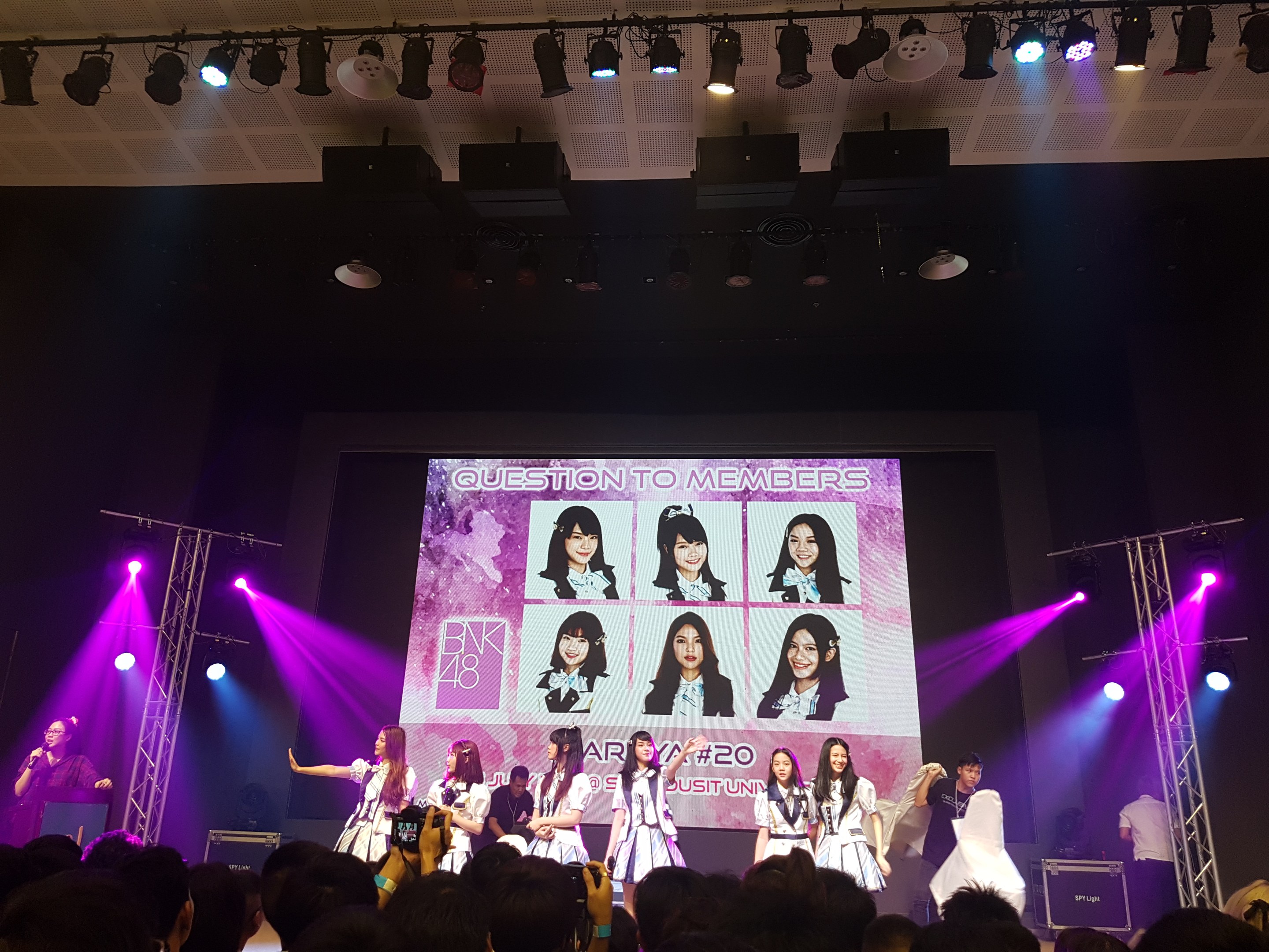 Eine Gruppe von Menschen, die auf einer hell erleuchteten Bühne performen, mit einer Menge darunter, die einen Sänger mit Mikrofon und einen Musiker zeigt, während ein Bildschirm im Hintergrund Bilder der K-Pop-Gruppe BK48 anzeigt.