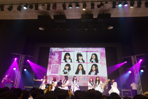 Eine Gruppe von Menschen, die auf einer hell erleuchteten Bühne performen, mit einer Menge darunter, die einen Sänger mit Mikrofon und einen Musiker zeigt, während ein Bildschirm im Hintergrund Bilder der K-Pop-Gruppe BK48 anzeigt.