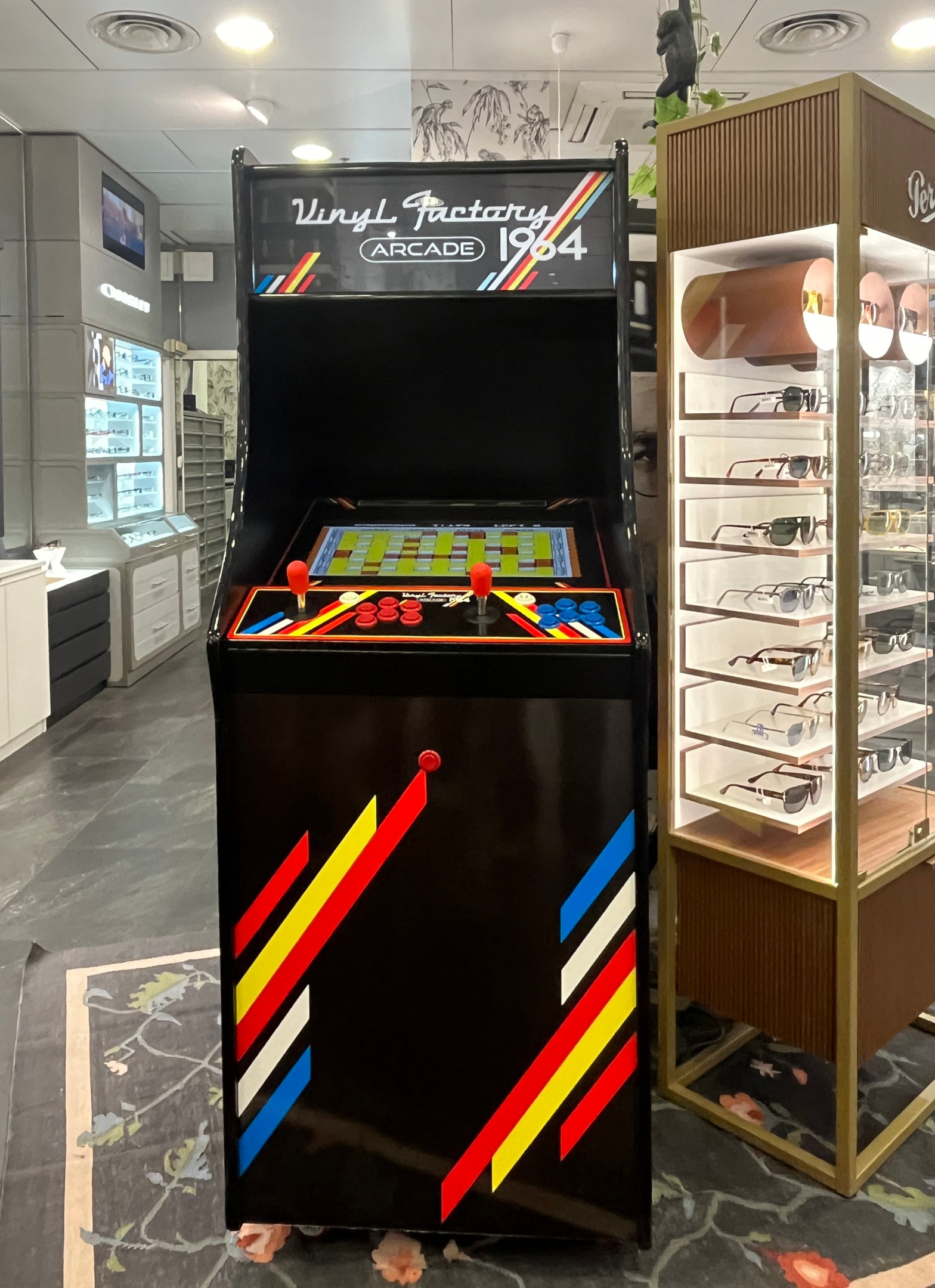 Ein Geschäft mit einem zentralen Retro-Arcade-Automaten, einem Regal mit Gläsern auf der rechten Seite und Hintergrund-Elementen wie Schränken, einem Fernseher und anderen Gegenständen, mit Deckenlampen, die eine retro-mäßige Atmosphäre schaffen.