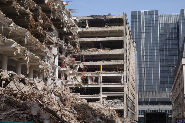 Ein gro├če Geb"ude in einer Stadt abgebrochen, umgeben von intakten Geb"uden, Stra├čeninfrastruktur, Fahrzeugen, B"umen und einem klaren Himmel.
