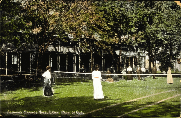 Altes Schwarz-Weiß-Foto einer Tennisveranstaltung auf dem Rasen des Abenakis Springs Hotel in Provo, Quebec, mit Spielern mit Schlägern und sitzenden Gästen auf einem Rasenplatz mit Netz, Bäumen und einem Gebäude im Hintergrund.