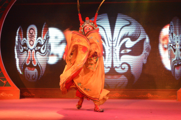 Eine Person in chinesischem Opernoutfit und Maske tritt auf der Bühne mit ausgestreckten Armen und zurückgelegtem Kopf auf, während ein Bildschirm im Hintergrund Bilder anzeigt.