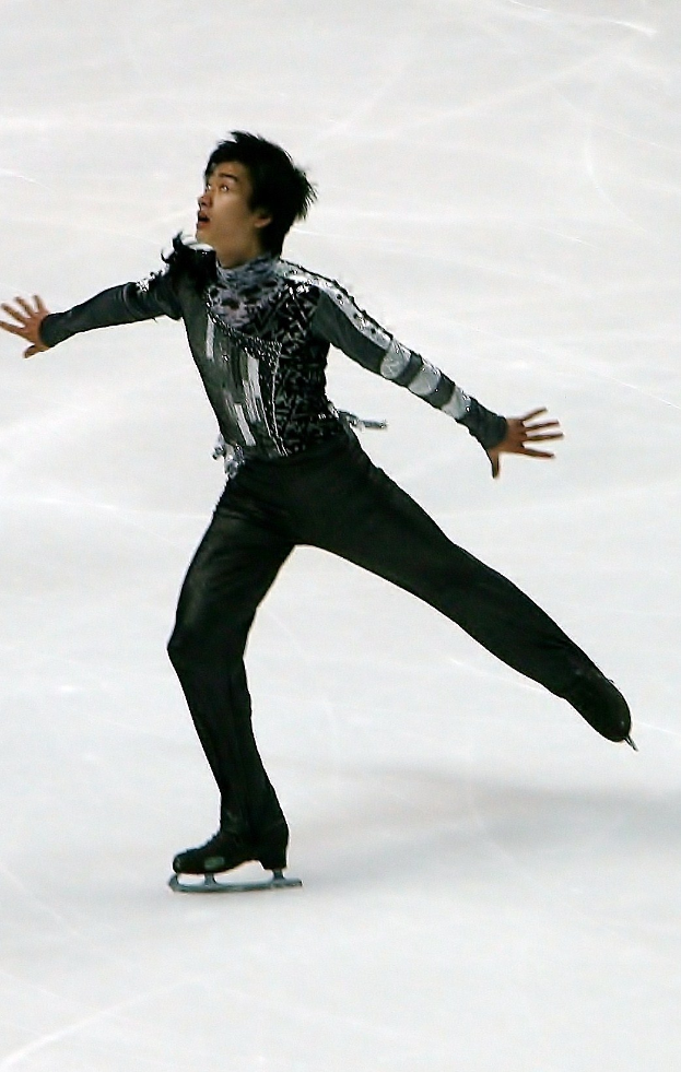 Männlicher Eiskunstläufer in einem schwarzen Kleid, der über eine Eisfläche gleitet und eine Choreographie mit synchronen Arm- und Beinbewegungen präsentiert.