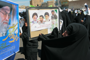 Eine Gruppe von Frauen in schwarzen Abayas hält Plakate mit Fotos von Ayatollah Ali Khamenei hoch, im Hintergrund sind Gebäude, Pfosten, Fahrzeuge und ein klarer blauer Himmel zu sehen.