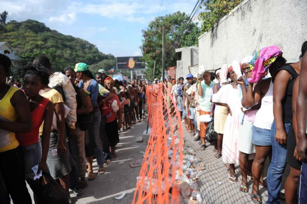 Lange Schlange von Menschen, die an einer Wahllokal-Schlange warten, mit einem Zaun, einer Wand, Bäumen, Gebäuden, Versorgungsmasten, Drähten und einer Werbetafel im Hintergrund.