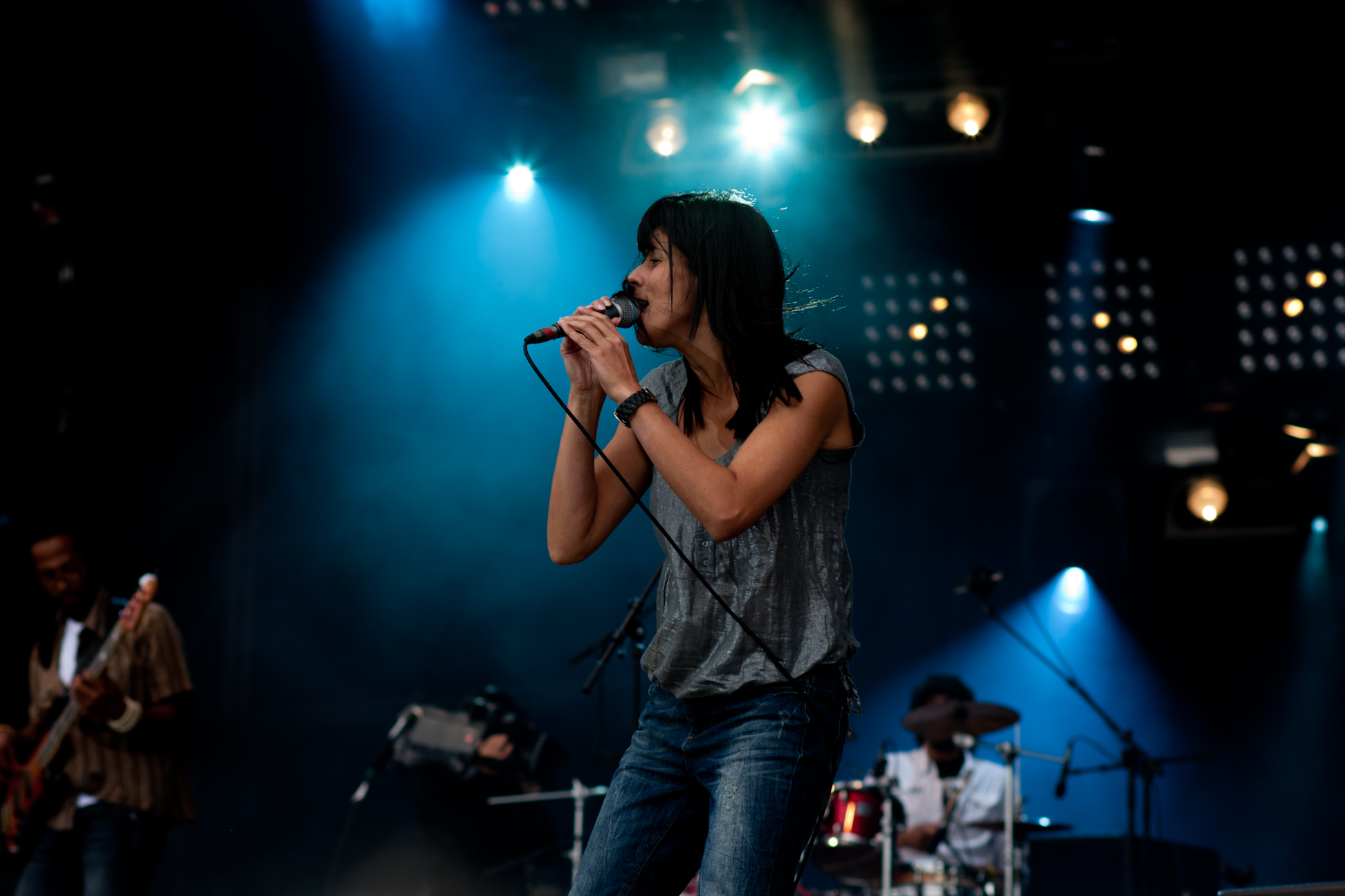 Eine Frau singt auf der Bühne in ein Mikrofon, flankiert von einem Mann mit Gitarre links und einer Person am Schlagzeug im Hintergrund, unter grellen Bühnenlichtern.