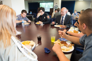 Eine Gruppe von Menschen sitzt an einem Tisch mit Tellern, Gläsern, Besteck und Servietten, mit Säulen, Vorhängen, einem Fenster und Deckenlampen im Hintergrund.