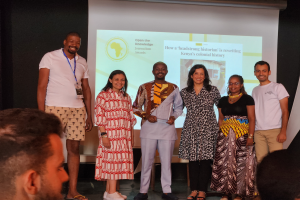 Eine Gruppe von Menschen auf einer Bühne, die fröhlich in die Kamera lächeln und posieren, mit einem Publikum davor und einem Bildschirm im Hintergrund, der 'Open the Knowledge Journalism Awards 2019' anzeigt.