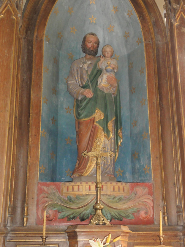 Statue of Jesus holding a baby in a church, with a cross symbol in the foreground, a wall in the background, and candles with stands and a flower vase at the bottom.