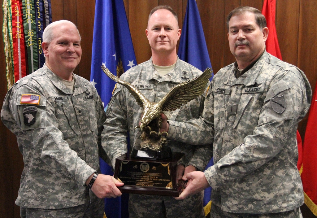 Drei Männer in Uniformen halten Auszeichnungen, mit Flaggen im Hintergrund und buntem Text in der linken oberen Ecke.