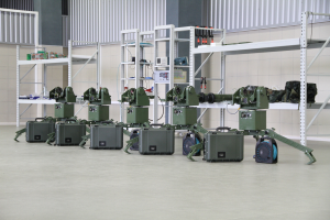 Eine Gruppe von Militärrobotern auf einem Boden, umgeben von Koffern und anderen Gegenständen auf Gestellen, mit einer Wand und einem Geländer im Hintergrund.